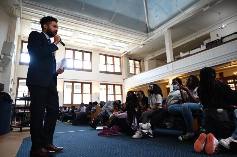 A young man in a suit speaks into a microphone while standing at the front of a church sanctuary. Teenagers sit in the audience.