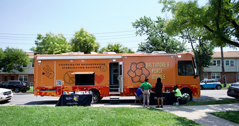 A large orange RV is parallel parked with a few people standing or sitting at a table in front of it.  