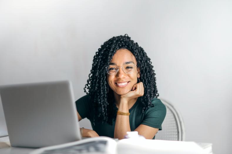 A young woman sits behind a laptop, smiling for the camera. 