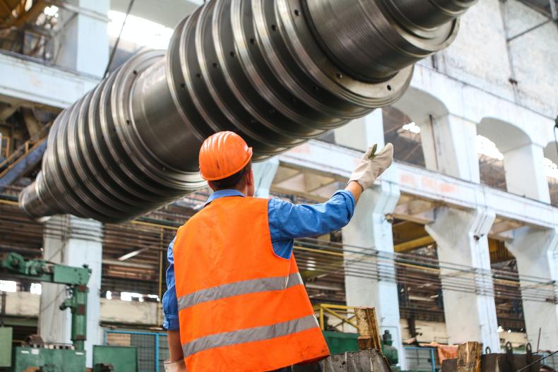 A worker in an orange vest and orange hard hat is seen from behind inside an industrial factory.