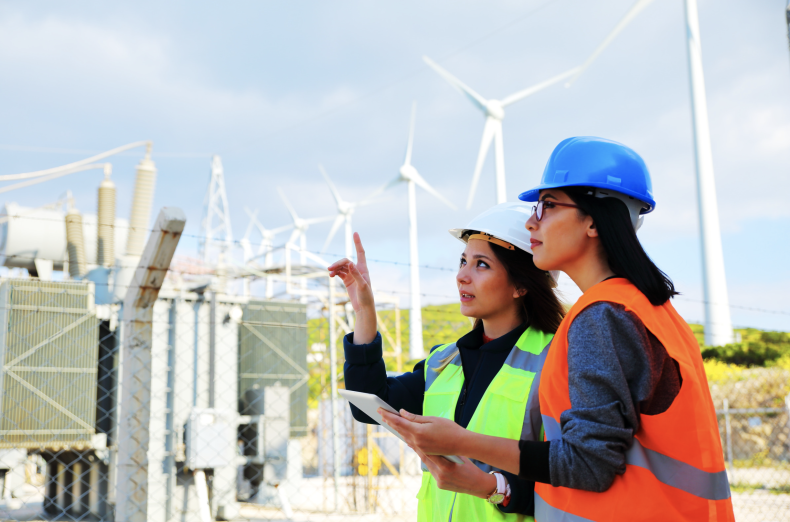 Two women in safety vests and hard hats stand outside among large white windmills.