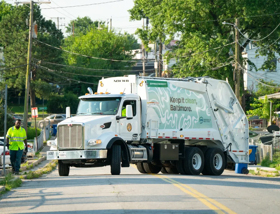 A large white garbage truck with "Keep it clean, Baltimore" written on the side is driving through a residential neighborhood. The truck has the Baltimore Department of Public Works (DPW) logo and features a green and white design with recycling arrows. A sanitation worker wearing a bright yellow safety vest walks alongside the truck, while trees and rowhouses line the street in the background.