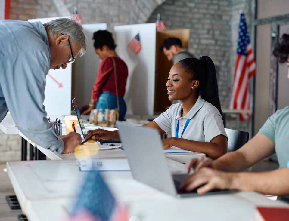 Senior US citizen signing up for voting with help of African American female volunteer at polling place.