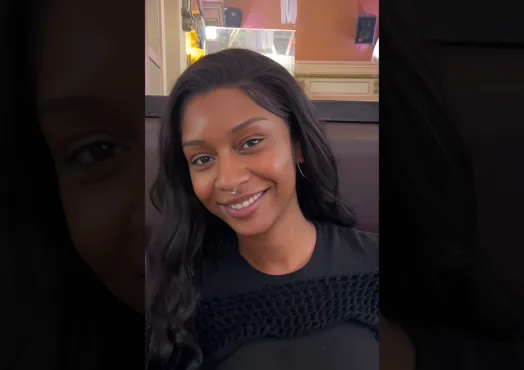 A young African American woman with long wavy hair and a septum piercing is wearing a black long sleeve top, smiling at the camera.