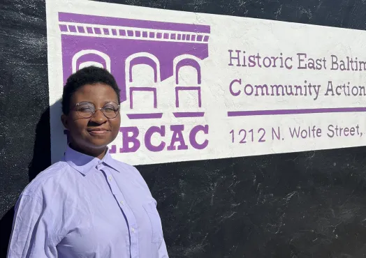 An African American young adult with a short haircut wearing circle glasses, a lilac button up shirt, and blue jeans outside of HEBCAC building