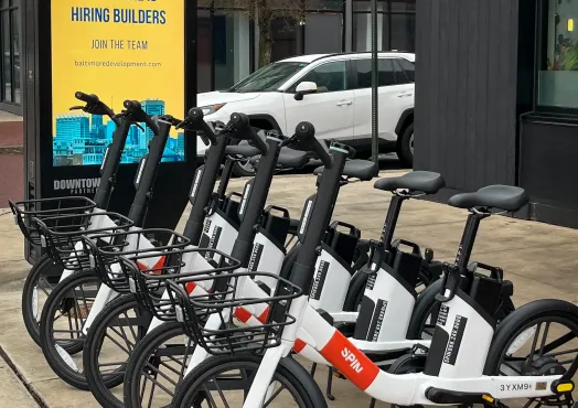 Photo of a row of Spin electric bikes parked at a docking station in Baltimore's Fells Point neighborhood. The white and black bikes with orange Spin branding are lined up on the sidewalk next to a digital advertising kiosk displaying a Baltimore Development Corporation recruitment ad that reads "Baltimore is Hiring Builders." A white SUV is visible in the background along with modern mixed-use buildings. The Baltimore City Department of Transportation logo appears in the lower right corner.