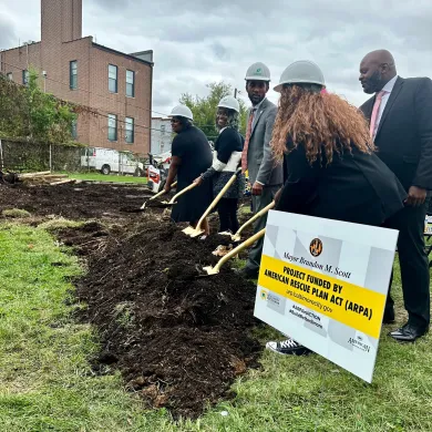 ARPA BCRP Playground Rebuild. A group of people in helmets participating in a groundbreaking ceremony with shovels. A sign in the foreground displays information about the project, indicating it is funded by the American Rescue Plan Act (ARPA). 