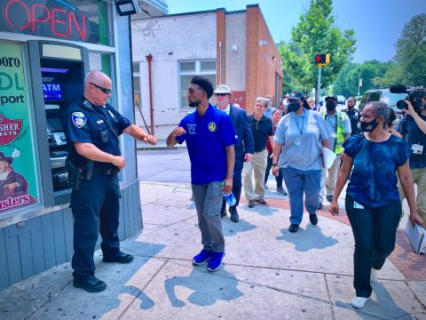 A crowd of people walks on a sidewalk and a young black man fist bumps a white police officer.