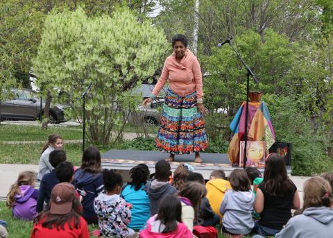A woman, who is standing, speaks to a small group of seated children in an outdoor setting.