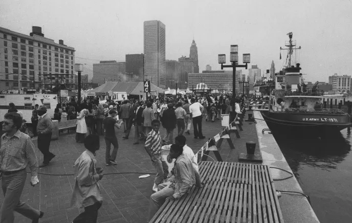 Black and white photograph from 1970s or 1980s of a crowd of people walking along the Inner Harbor promenade with water on the right hand side where a tub boat is docked. The perspective looks north towards the business center of the City.