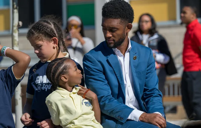 Mayor Brandon M. Scott, in a blue suit, is sitting on a bench outdoors during the Cecil Playground Ribbon Cutting event, engaging warmly with a young child. One child, in a pale yellow shirt and braided hair, leans against him, looking up attentively. The other child, with curly hair and a dark blue shirt, stands nearby, observing. In the blurred background, several people, including someone in a red shirt, are visible against a building with colorful panels.
