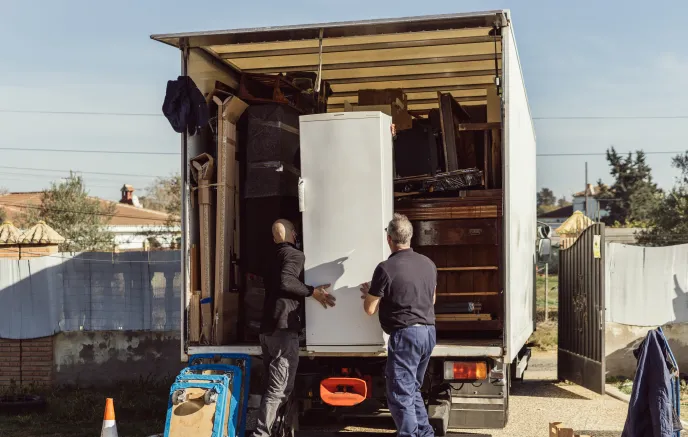 Two workers are loading a large white refrigerator onto a moving truck filled with various household items, including furniture and boxes. The setting is an outdoor area with a fence and a residential neighborhood in the background. One worker is wearing a black jacket and mask, while the other is dressed in a dark blue polo shirt and blue pants. A traffic cone and moving equipment are visible near the truck.