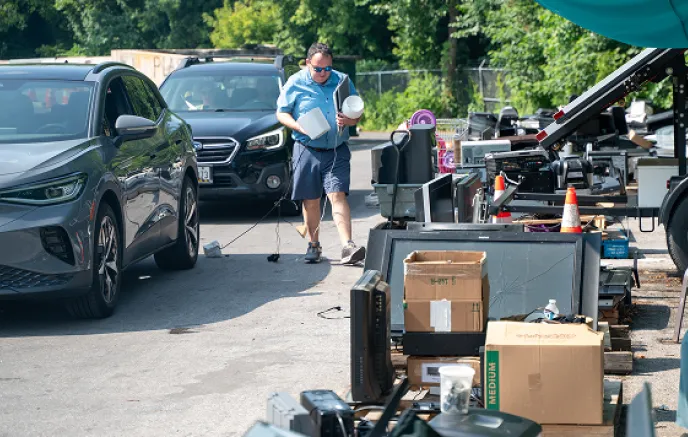  A man wearing sunglasses, a blue short-sleeve shirt, and shorts is walking toward a designated e-waste collection area, carrying old electronics, including a laptop and a lamp. Several cars are parked nearby, with other community members participating in the drop-off event. The area is filled with discarded televisions, monitors, printers, and other electronic devices, neatly placed on pallets or in bins for sorting. Orange traffic cones and a conveyor belt system are visible in the background, suggesting 