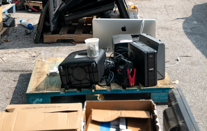 A wooden pallet loaded with discarded electronic devices is placed on the ground at an electronics recycling event. The pile includes an old iMac computer, a black printer, an uninterruptible power supply (UPS) unit, tangled cords, and miscellaneous electronic accessories. Nearby, open cardboard boxes contain additional electronic waste. In the background, more pallets with stacked electronic devices, including televisions and monitors, are visible, indicating a dedicated e-waste drop-off area. The setting 