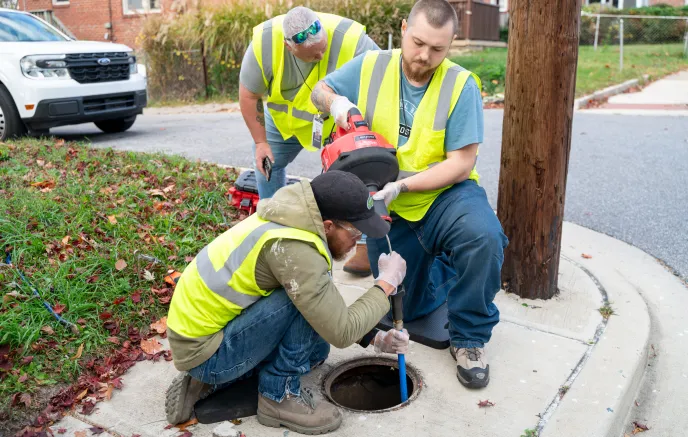 Three utility workers wearing high-visibility safety vests conduct maintenance at an open manhole on a sidewalk. One worker is crouching and using a testing device, while another holds equipment. A third worker observes while standing nearby. A white pickup truck is parked in the background near a grassy area with fallen leaves.