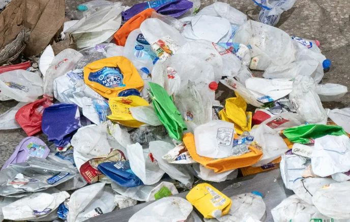 A pile of discarded plastic containers, including detergent bottles, milk jugs, and other rigid plastic packaging, scattered on the ground. Some containers appear dirty and partially crushed