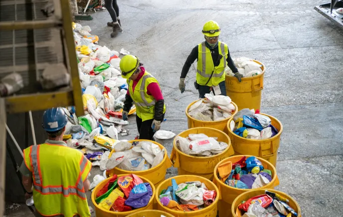 Workers in high-visibility vests and hard hats sorting plastic waste in an industrial recycling facility. Large yellow bins filled with plastic waste are lined up, while more discarded plastics are spread on the floor