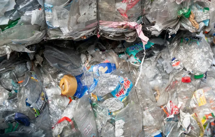 Stacks of tightly compressed plastic waste bales are piled up outdoors, consisting of crushed plastic bottles, containers, and other recyclable materials. Various colors, labels, and brands are visible among the plastic debris. The setting appears to be a recycling or waste management facility, where plastic waste is collected and prepared for processing. In the background, there are trees and an open sky, indicating that the facility is in an outdoor or semi-outdoor environment.