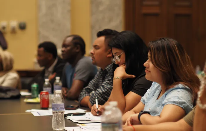 Several people sitting at a long table, taking notes and listening