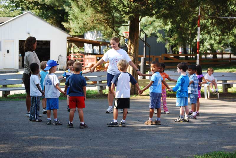 Children in a circle play a game at a Camp Olympia, guided by an adult in a sunny outdoor setting.