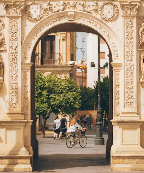 View through an archway of a girl on a bicycle with people walking on the street