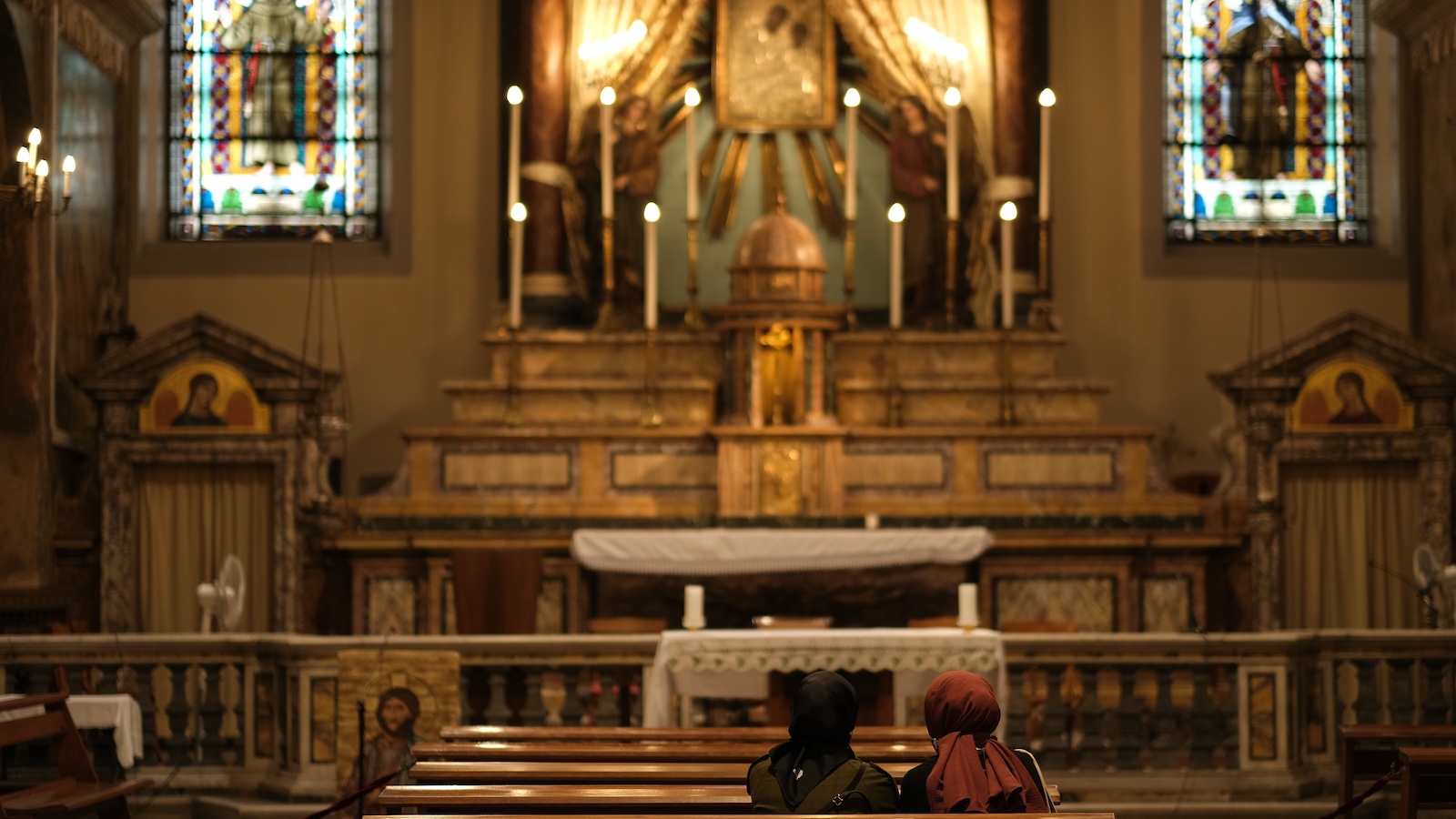 Two women wearing head coverings sit in a Church surrounded by candles