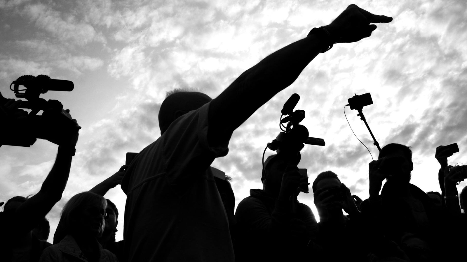 Silhouette of a man pointing surrounded by people with cameras underneath a cloudy sky