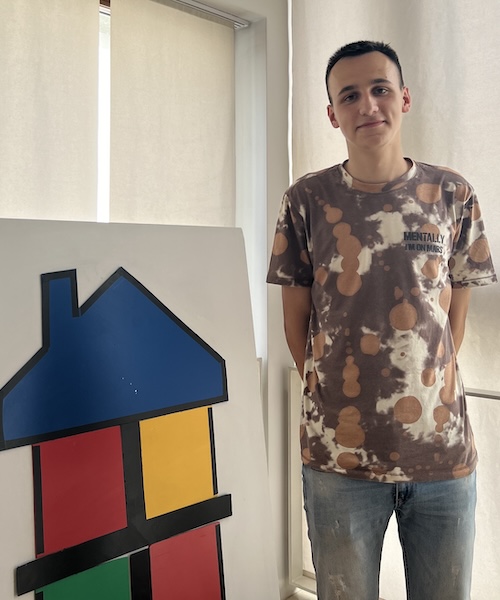 Manuel standing in a brown shirt and jeans next to a drawing of a house
