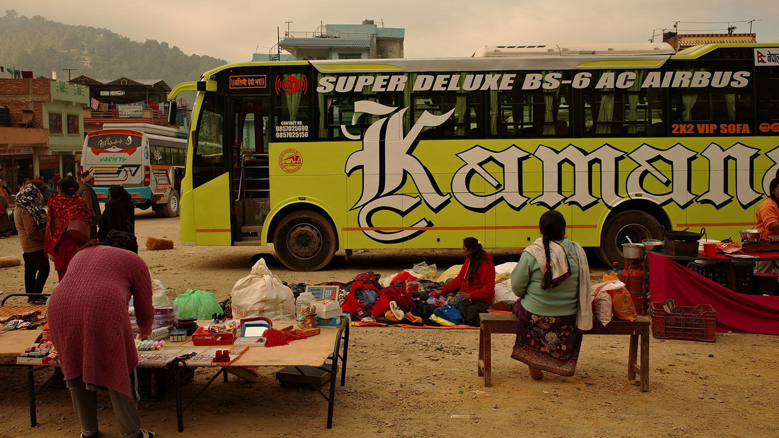 A bright yellow bus with "Super Deluxe" signage in the middle of female vendors in an outdoor market in Nepal