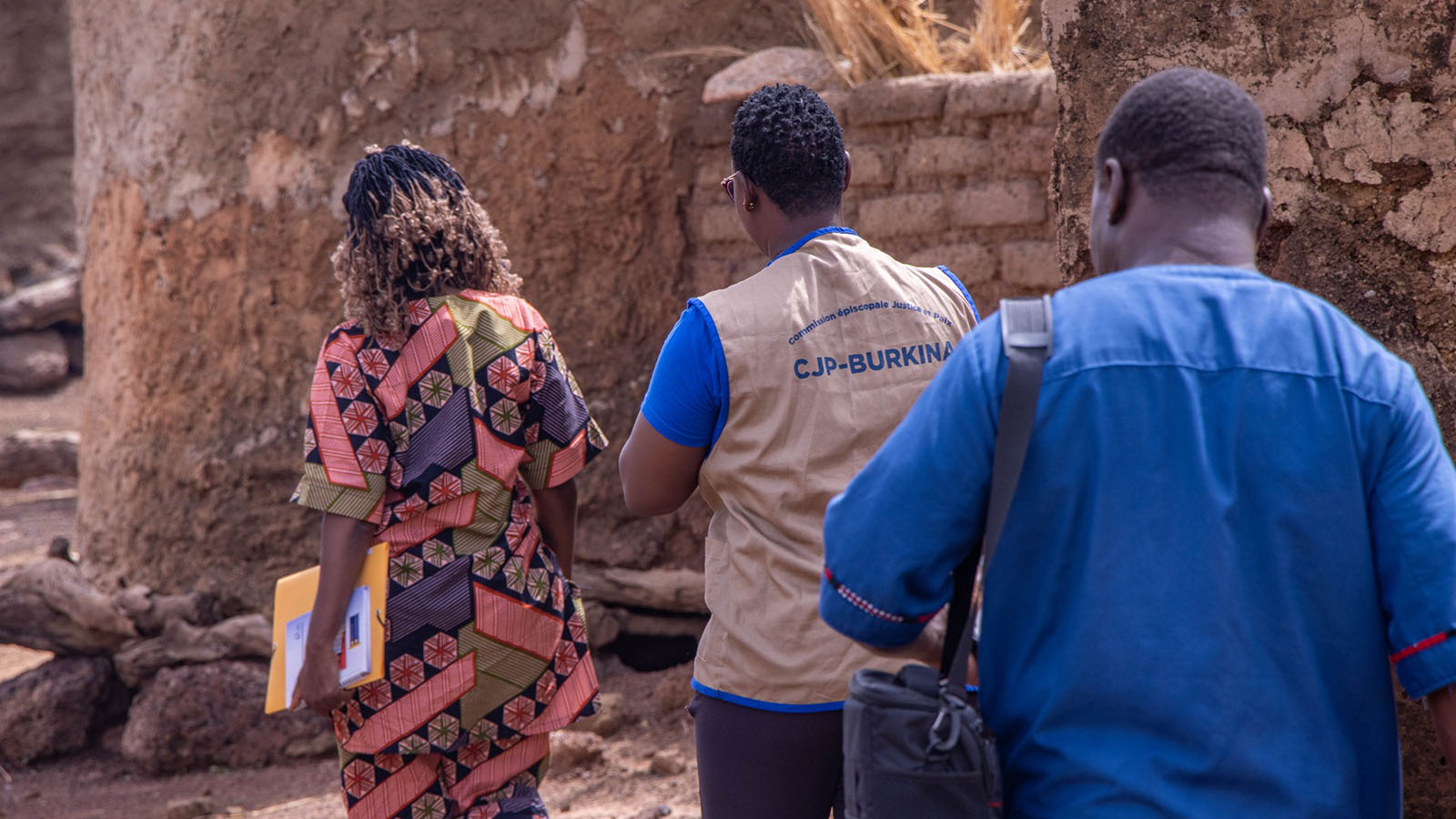 Three people walking in front of mud rick walls, with one wearing a CJP-Burkina vest.