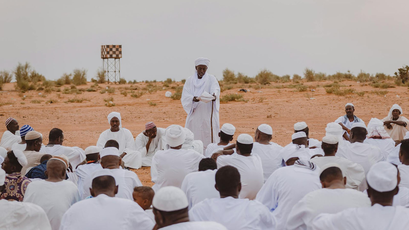 Men in white robes sitting outside in the desert, listening to another man speaking