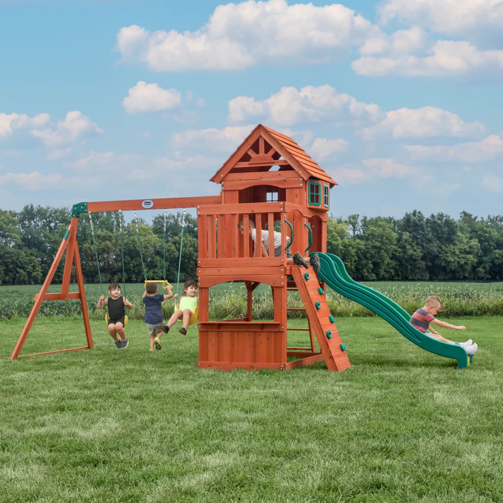 A group of children happily play on the cedar swings and slide in the backyard. Trees can be seen in the background, against a backdrop of a cloud-like sky.