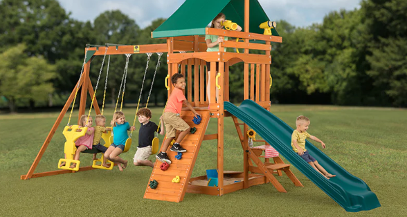 A group of children played on a cedar playsets and a cedar swing sets in the backyard. Trees can be seen in the background, against a cloud-like sky.