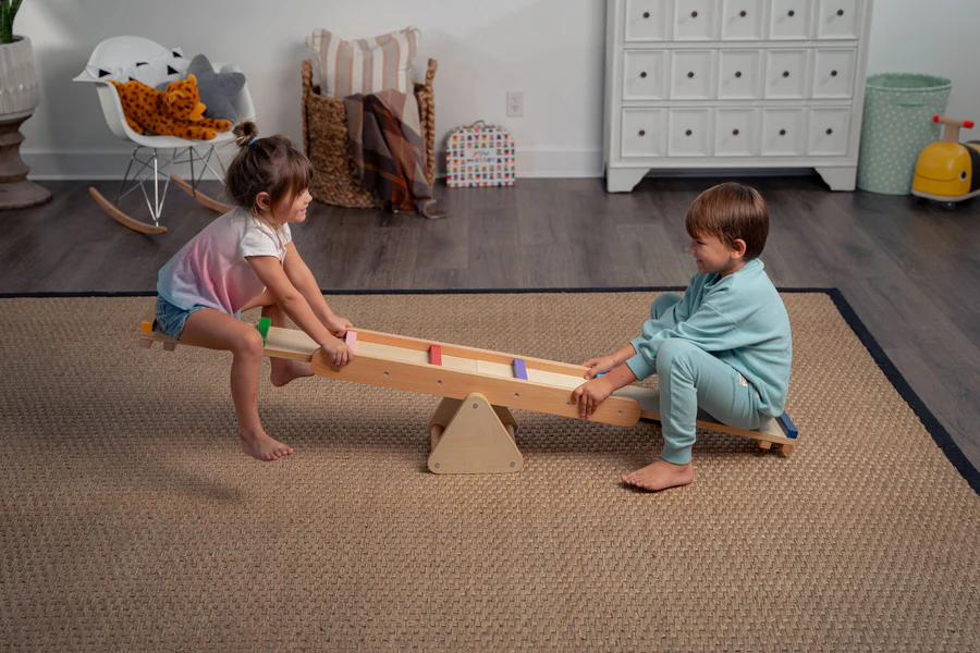 Little boy and little girl playing on wooden indoor seesaw in empty space at home
