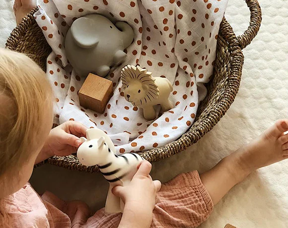 Little Girl Playing With Tikiri Natural Rubber Teethers In A Bamboo Basket On The Ground