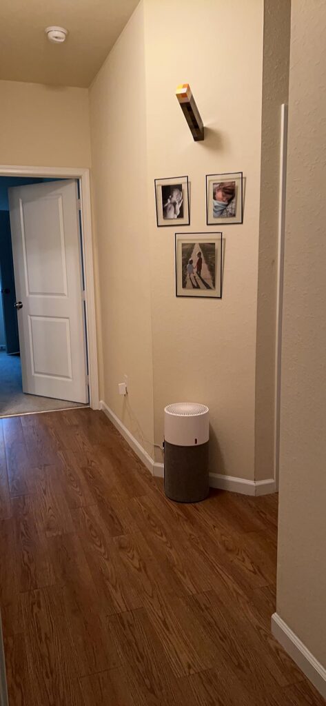 A bedside air purifier placed on a wooden nightstand in a calm bedroom setting with soft neutral tones and decorative vase.