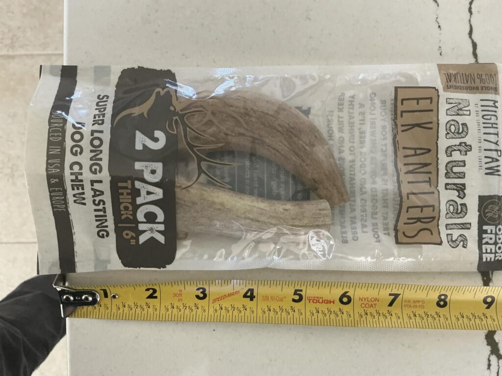 Close-up photograph of a clear plastic bag containing two natural elk antlers, part of the Mighty Paw Naturals line. Product text includes: 'ELK ANTLERS', '2 PACK THICK | 6"', 'SUPER LONG LASTING DOG CHEW', 'SOURCED IN USA & EUROPE', 'ODOR FREE', 'SINGLE INGREDIENT', and '100% NATURAL'. A yellow tape measure is placed below the bag, confirming the length is over 6 inches.