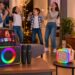 A lively family night scene where parents and their two children are jumping, dancing, and singing in a warm living room with a large TV. In the foreground, the black horizontal Peski karaoke machine (with tablet) sits on a side table, while the rose gold mini karaoke machine and the tall tower speaker with flashing RGB lights are placed on separate tables around the room, creating an immersive party environment.