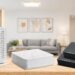 A studio product showcase of three Hitron devices side-by-side on a wooden surface against a blurred warm living room background: a vertical white modem, a horizontal white modem, and a stacked pair of black MoCA adapters.