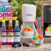 A vibrant brand lifestyle image from Hawaiian Shaved Ice titled "Bring the Aloha Home." A diverse product lineup is set up for an outdoor family party, including a white S900A and a black S777 shaved ice machine, three popular syrup flavors (Grape, Cherry, Blue Raspberry), colorful cups, spoon straws, and various prepared treats. Limes, strawberries, and drinks are on the table. A family is enjoying themselves in the background.