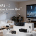 A scene in a modern and comfortable living room serving as the ELEPHAS Home Cinema Hub. A large screen displays an astronaut on the moon. Various ELEPHAS projectors (including black and white models, some on mini tripods, one in a hand) are arranged on marble platforms in the foreground. Top text features "ELEPHAS - Your Home Cinema Hub", and four icons with text detail features: "1080P HD & 4K Support", "Hi-Fi Stereo Sound", "WiFi & Bluetooth Connectivity", "Portable for All Occasions".