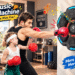 "Promotional 16:9 hero image of the OurStarry Smart Music Boxing Machine mounted on a wall in a realistic home-gym setting, showing an adult and a child wearing boxing gloves actively punching the black hexagonal LED target. Bright English text annotations and stylized cartoon graphic markers highlight key selling points such as 9 training modes, Bluetooth music sync, and LED light targets."