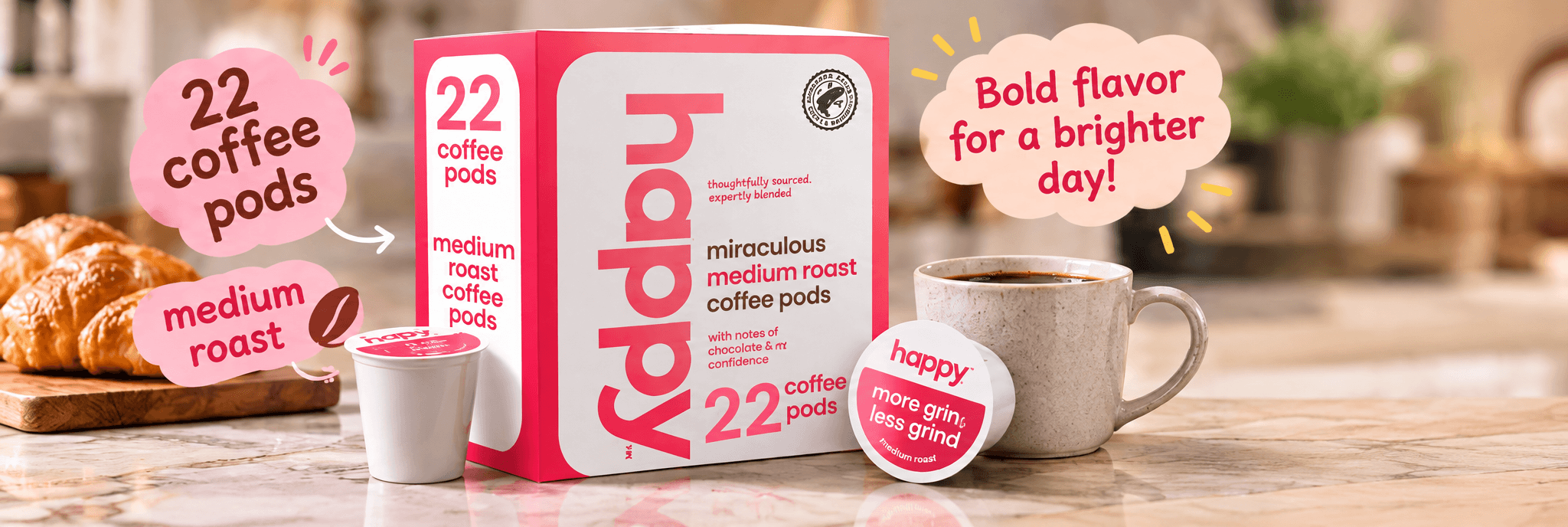 Alt (EN) Pink happy coffee-pod gift box printed with "22 coffee pods", "miraculous medium roast coffee pods", "thoughtfully sourced, expertly blended", "with notes of marshmallow and comfort" and "more grin, less grind". Hand-drawn callouts read "22 coffee pods", "medium roast" and "Bold flavor for a brighter day!". A single pink-lidded pod, an espresso cup and a stoneware mug of black coffee sit on a marble counter, with croissants on a wooden board at left and a softly blurred warm kitchen behind.