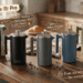 A warm, sunlit editorial shot of the full SterlingPro French press collection โ polished silver, matte black, grey, dusty blue and olive green โ arranged in a row on a walnut kitchen counter with steaming coffee, fresh croissant and scattered coffee beans, overlaid with playful hand-lettered English annotations: "Brew Like a Pro", "Double-Walled โ Coffee stays HOT for hours", "8000+ Happy Brewers" and "From our kitchen to yours โ the morning ritual, elevated".