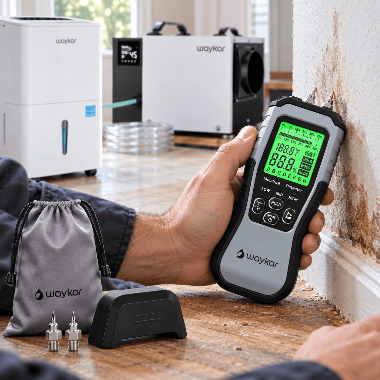 Waykar digital moisture meter with a green backlit LCD and dual metal probes, held against a damp interior wall during inspection, with testing accessories and a storage pouch nearby.