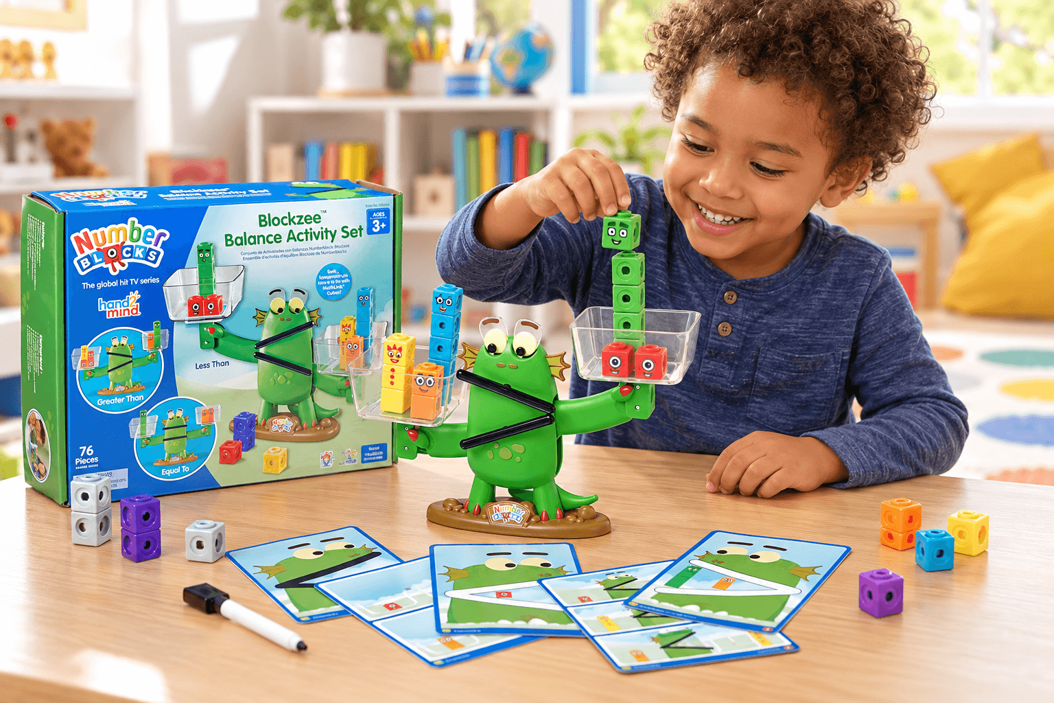 A smiling boy plays with the Numberblocks Blockzee Balance Activity Set at a wooden table. The green balance character holds colorful counting cubes in clear trays, with activity cards and the original product box nearby.