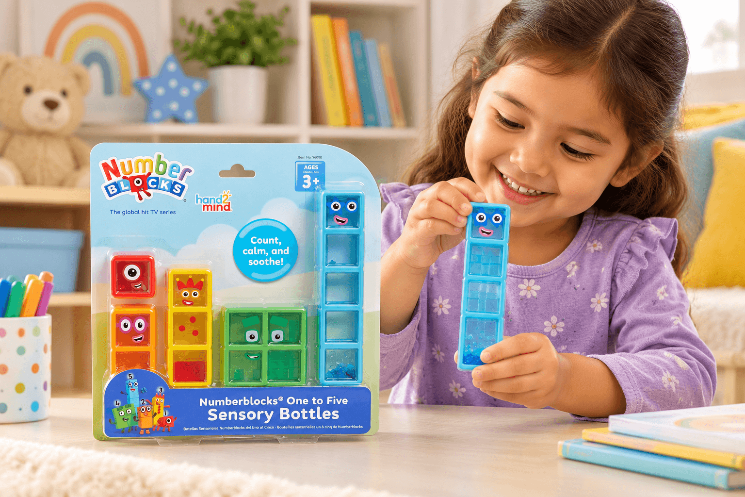 A smiling girl holds a blue Numberblocks Five sensory bottle in a cozy playroom. The Numberblocks One to Five Sensory Bottles package sits beside her, displaying colorful number characters from one through five.