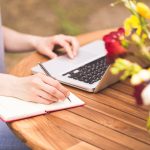 Close-up on woman hands writing on notebook and learning in the garden with bunch of colourful flowers in front of her. Freelancer working in the garden. Writing, surfing in the internet. Young girl. Relax and joy.