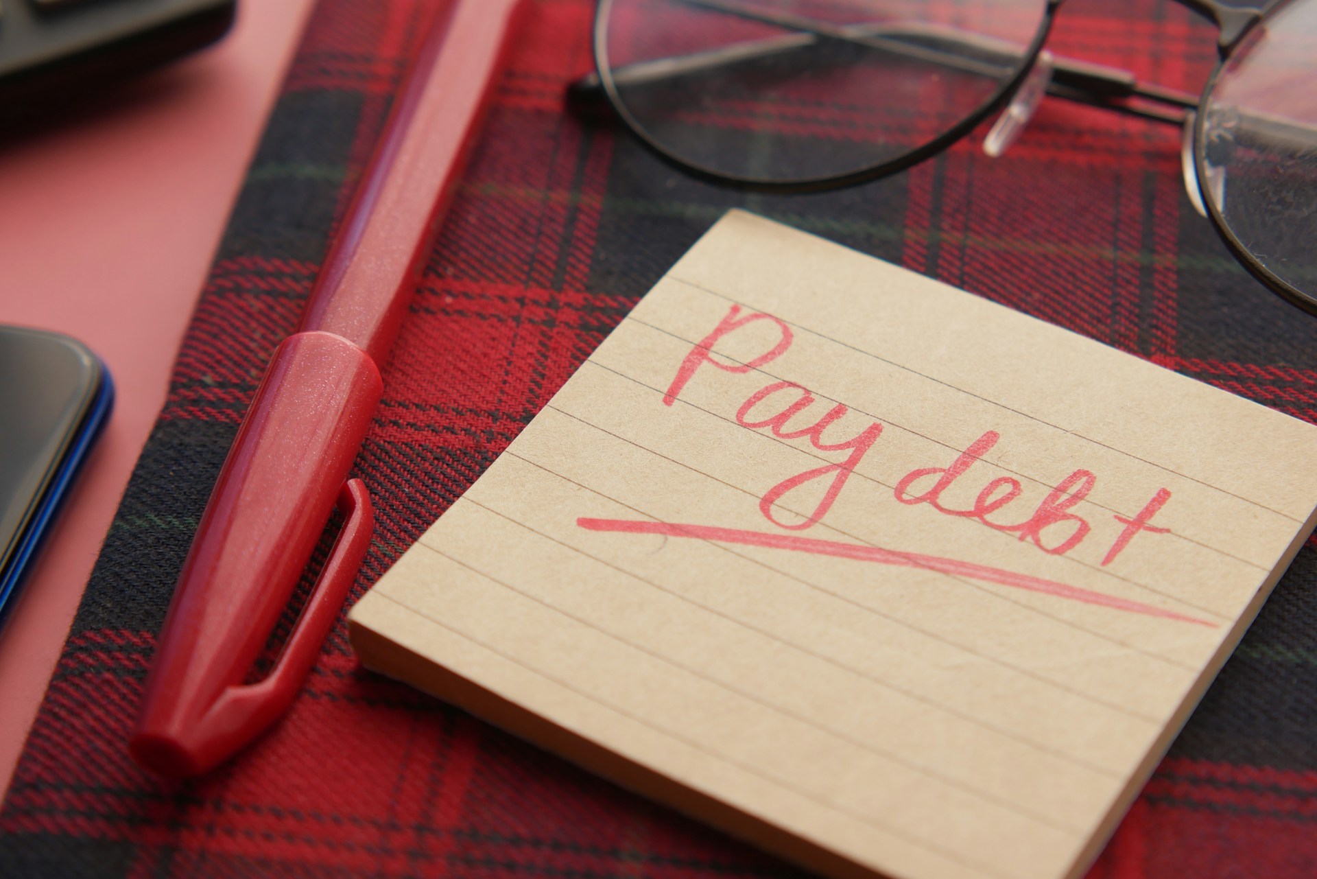 A note that says pay debt next to a pen and glasses