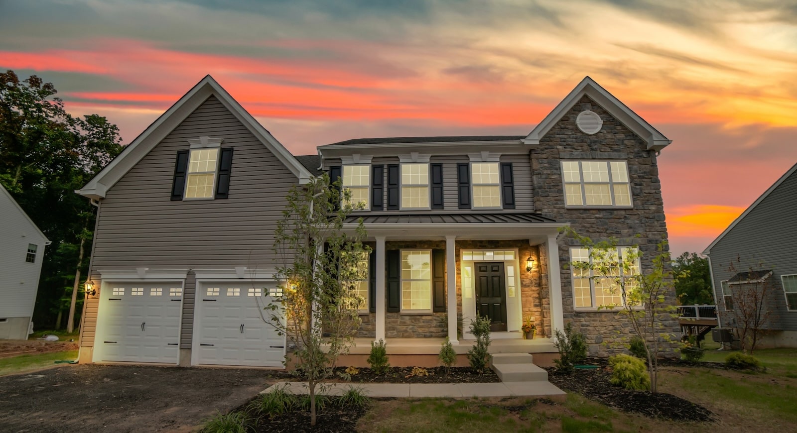 Two-story suburban home with stone and siding exterior, black shutters, and a double garage, photographed at sunset with a vibrant pink and orange sky.